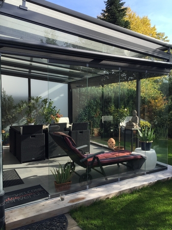 Moderner Wintergarten mit Glaswänden, Loungemöbeln und Pflanzen im sonnigen Garten.